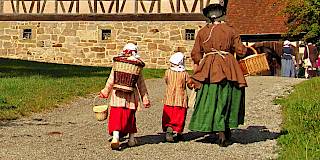 Eine Person in mittelalterliche Kleidung und einem Korb geht mit zwei ebenfalls historisch gekleideten und Körben ausgestatteten Kindern zu einem großen Fachwerkhaus