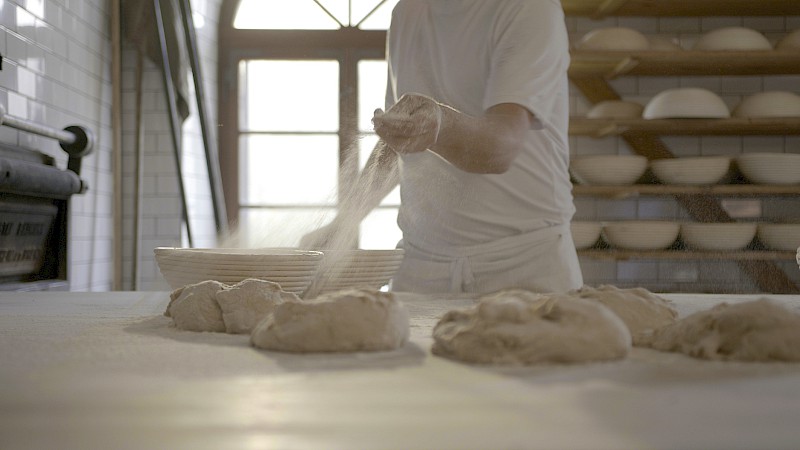 Ein Bäcker streut Mehl über ungebackene Brotlaibe, im Hintergrund Regale mit leeren Brotfomen