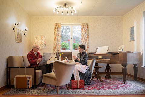 Ein älteres Ehepaar sitzt gemeinsam in ihrem fünfziger Jahre Wohnzimmer. Sie sitzt auf einem Sessel und flickt ein Hemd, er sitzt ihr gegenüber auf der Couch und liest Zeitung