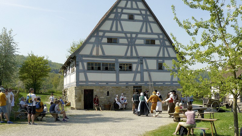 Nachgestellte historische Dorfszene: Tanzende Menschen in Tracht vor einem viergeschossigen Fachwerkhaus, Besucher:innen schauen der Gruppe zu