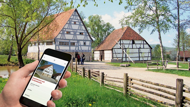 Zwei Fachwerkhäuser nebeneinander, im Vordergrund eine Hand mit Smartphone, darauf erläutert die Museumsapp, dass es sich bei einem Gebäude um das Bauernhaus aus Zaisenhausen handelt.