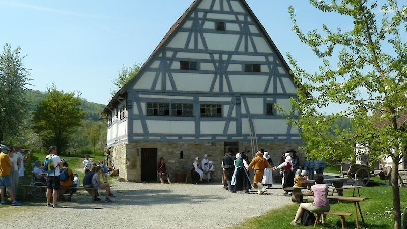 Ein altes Bauernhaus im Fachwerkstil, davor historisch gekleidete Menschen gemeinsam tanzend, drum herum schauen Besucher:innen der Festgemeinde zu